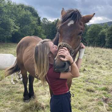 A young woman lovingly hugs and kisses a brown horse in a grassy field, while a white horse grazes nearby. Trees and cloudy sky surround them.