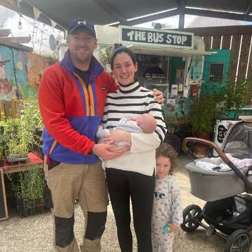 A smiling family of four poses outside a quirky food stand called "The Bus Stop." The mother holds a newborn, while a toddler stands nearby, stroller beside them.