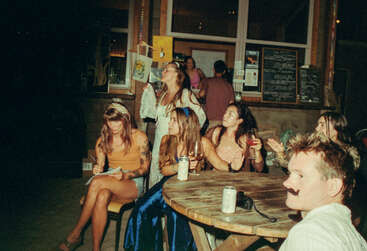 A group of people in costumes sit and stand around a wooden table, some with drinks. The atmosphere is festive, casual, and possibly part of a themed event.