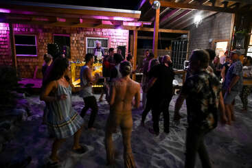 A lively group of people dance barefoot on sandy ground at a nighttime outdoor party, illuminated by purple and yellow lights, with a DJ playing music.