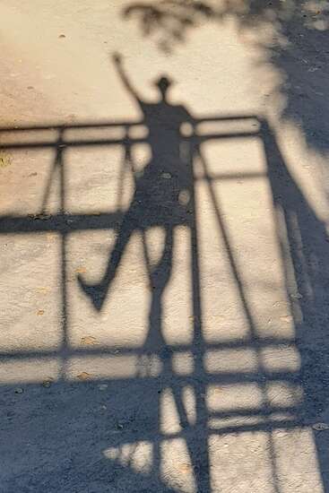 La sombra nítida de una persona de pie en un puente, saludando o levantando un brazo, se proyecta sobre el suelo bajo la brillante luz del sol. Las líneas geométricas se cruzan.