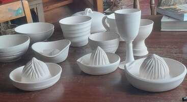 Esta imagen muestra una mesa de madera con varias piezas de cerámica sin esmaltar, entre las que se incluyen cuencos, tazas, exprimidores de cítricos, una copa y una fuente con tapa. Un espacio de trabajo acogedor y creativo.