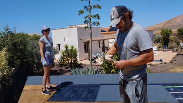 A man and woman in casual clothing work outdoors, possibly on a roof, with a house, plants, and clear blue sky in the background under the sun.