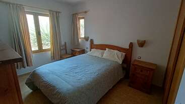 This bedroom features a wooden bed and side tables, blue bedspread, two windows with light curtains, wooden chair, minimal decor, and bright natural light.