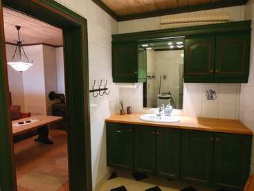 Une salle de bains au style rustique, avec des meubles verts, un plan de travail en bois et un grand miroir. On aperçoit la pièce voisine, où se trouvent une table en bois, des chaises, un luminaire et une décoration chaleureuse.