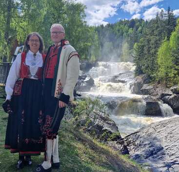 Una pareja sonriente, vestida con trajes tradicionales noruegos, posa junto a una pintoresca cascada, rodeada de frondosos árboles bajo un cielo primaveral luminoso y parcialmente nublado.