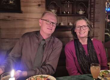 Un hombre y una mujer están sentados juntos en un acogedor restaurante de madera. Sonríen con calidez, van bien vestidos y tienen platos de comida y bebidas sobre la mesa que tienen delante.