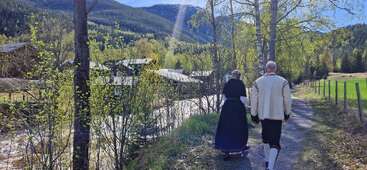 Un hombre y una mujer vestidos con trajes tradicionales caminan por un sendero junto al río. La escena está rodeada de árboles primaverales, montañas y edificios agrícolas bajo la luz del sol. Ambiente tranquilo.