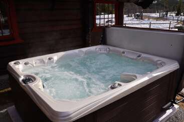 Junto a una rústica cabaña de madera hay un jacuzzi humeante. La nieve cubre el jardín y las colinas de los alrededores, creando un acogedor contraste con el agua caliente que burbujea.