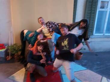 Six people pose in a playful, chaotic manner, some stacked atop each other, one playing a recorder. The scene is lively, fun, and slightly blurry.