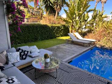 Un elegante patio exterior cuenta con un acogedor sofá con cojines decorativos, una mesa de cristal, una exuberante vegetación, flores de vivos colores, una terraza de madera, tumbonas y una piscina cristalina.