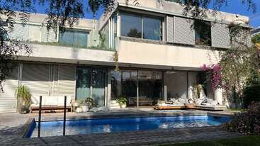 Una moderna casa de dos plantas con amplios ventanales da a una piscina azul. La vegetación circundante, las zonas de descanso al aire libre y los acogedores rincones para relajarse crean un ambiente relajante.