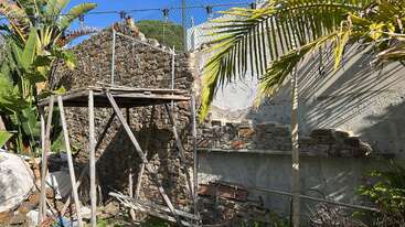 Esta imagen muestra un muro rústico de piedra en construcción, rodeado de plantas tropicales y un andamio de madera improvisado, con la luz del sol proyectando sombras sobre la estructura sin terminar.
