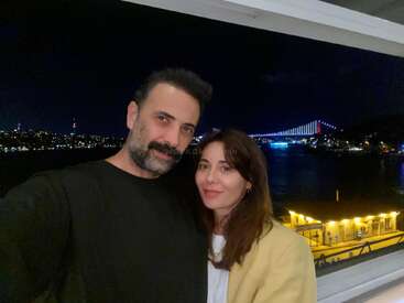 Una pareja posa para hacerse un selfi por la noche con un puente bellamente iluminado y el perfil de la ciudad al fondo, junto a una zona ribereña que resplandece con las luces.