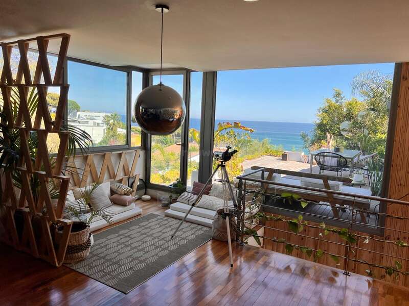 Un espacio habitable moderno, con ventanales de suelo a techo, cómodos asientos, plantas y un telescopio, ofrece unas preciosas vistas al mar, combinando el confort del interior con la naturaleza.