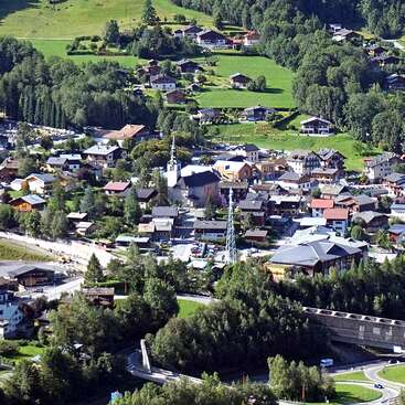 La imagen representa una pequeña ciudad serena enclavada en las montañas, con un campanario de iglesia, casas, árboles y una carretera, con un puente en primer plano.
