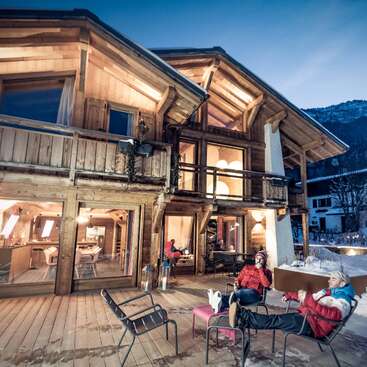La imagen representa un sereno chalet de madera con una amplia terraza, donde dos personas se relajan en sillas, rodeadas de montañas nevadas y un cielo azul despejado al fondo.