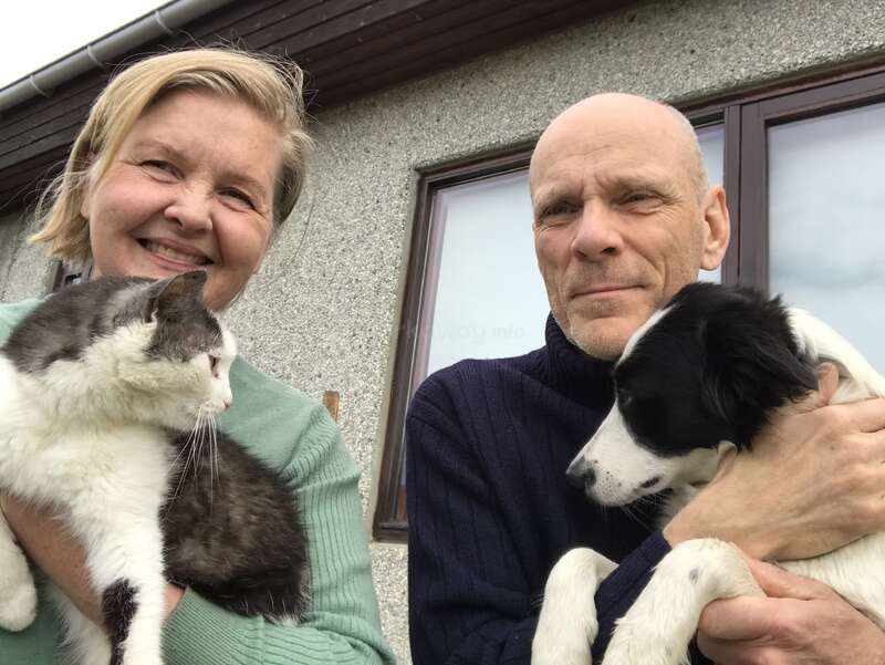 Uma mulher sorridente segura um gato, enquanto um homem segura um cachorro. Eles estão do lado de fora de uma casa, demonstrando carinho e afeto pelos seus queridos animais de estimação.