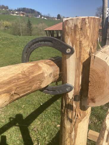 Uma ferradura é usada de forma criativa como um suporte para unir dois troncos de madeira em uma cerca rústica ao ar livre, com grama verde e o campo ao fundo.