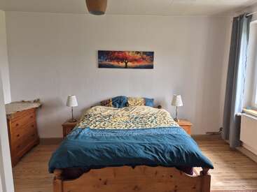 A cozy bedroom with a wooden bed, blue and yellow bedding, two bedside tables with lamps, a dresser, and a colorful tree painting above the bed.