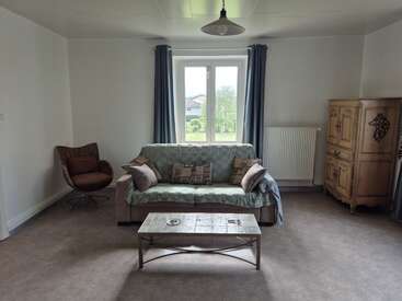 A cozy living room with a sofa, multiple cushions, coffee table, wicker chair, wooden cabinet, large window with blue curtains, and calming, neutral-toned decor.