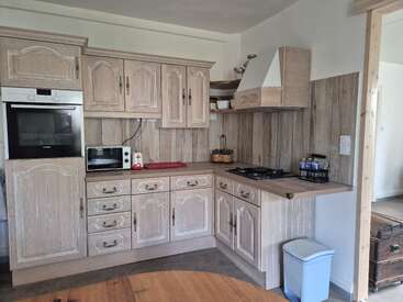 This image shows a rustic-style kitchen with light wood cabinets, built-in oven, microwave, gas stove, countertop essentials, a small trash bin, and wood-paneled walls.