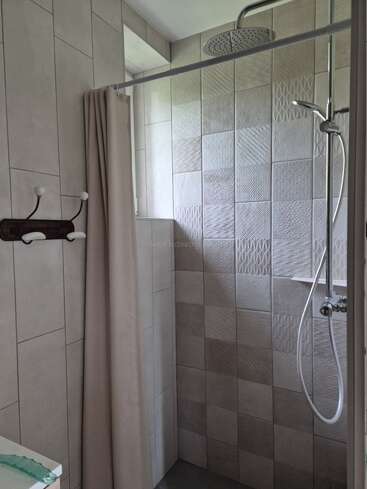 This image shows a modern shower area with light gray textured tiles, a rainfall showerhead, a handheld shower, a beige curtain, and a wall-mounted coat rack.