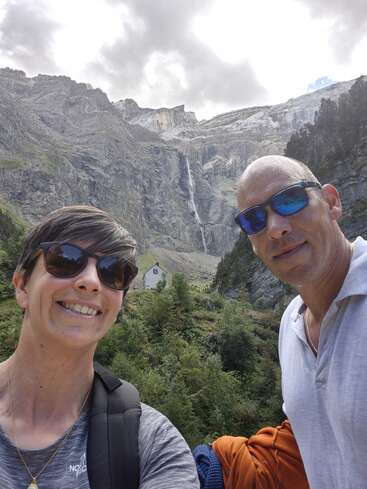 A smiling couple wearing sunglasses takes a selfie outdoors, with a mountainous landscape, greenery, a small house, and a tall waterfall in the background.
