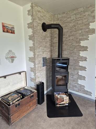 In einer gemütlichen Ecke stehen ein moderner schwarzer Holzofen mit einem Rohr, eine Ziegeltapete, eine alte Truhe mit Brettspielen und ein Wandregal.