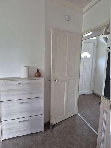 Ein einfaches Zimmer mit einer hellen Holzkommode mit einer Vase und kleinen Gegenständen, einer angelehnten weißen Tür und einem hellen Flur mit einer weißen Eingangstür.