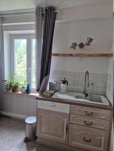 Eine helle Küche mit einem großen Fenster, Topfpflanzen, weißer Spüle, Holzschränken, Geschirrspüler, Seifenspender, grauen Vorhängen, Wandleuchten und einem Mülleimer.