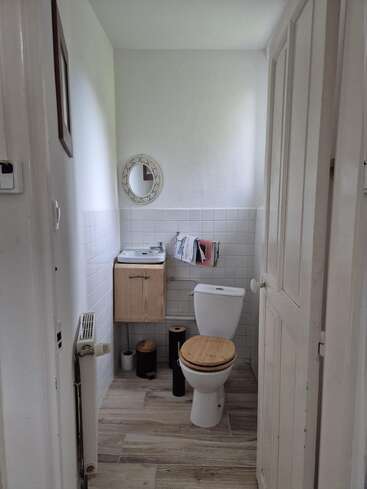 Dieses Bild zeigt ein kleines, aufgeräumtes Badezimmer mit einer Toilette, einem Toilettensitz aus Holz, einem kompakten Waschbecken, einem runden Spiegel, Lesestoff und hellem Holzdekor. Einfach und ordentlich.