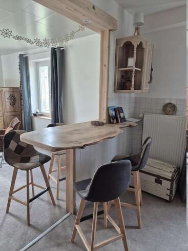 Dieser gemütliche Raum verfügt über eine hölzerne Frühstückstheke mit drei Hockern, einen Hängeschrank, Bücher und dekorative Elemente. Natürliches Licht und neutrale Töne schaffen eine einladende Atmosphäre.