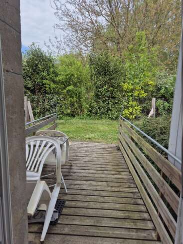 Eine Holzterrasse führt zu einem grasbewachsenen Hinterhof mit üppigen grünen Büschen und Bäumen. Auf der Terrasse stehen weiße Plastikstühle und ein kleiner Tisch.