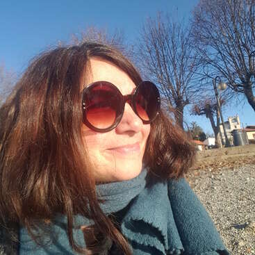 A woman with long brown hair and large sunglasses smiles outdoors on a sunny day. She wears a teal scarf. Bare trees and blue sky are visible.