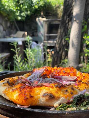 Eine frisch gebackene Pizza mit roten Zwiebeln liegt in einer gusseisernen Pfanne draußen, umgeben von viel Grün und natürlichem Sonnenlicht, was eine rustikale, einladende Atmosphäre schafft.