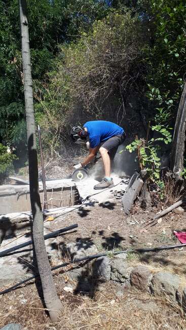 A person wearing a blue shirt, shorts, and safety gear uses a power saw to cut concrete outdoors, surrounded by trees, dirt, and construction materials.