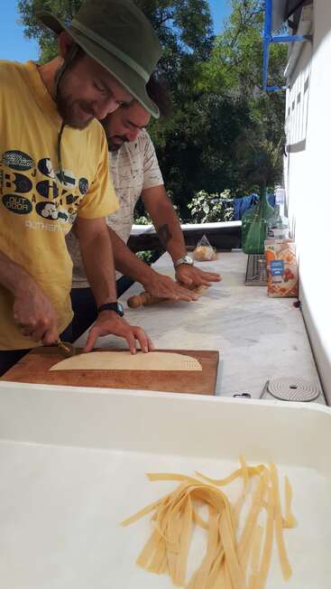 Two men outdoors are making fresh pasta. One is rolling dough, the other cutting it. Sunlight, green trees, juice box, and pasta strips complete the scene.
