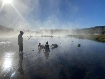 Quatro pessoas relaxam em uma fonte termal enevoada sob um sol brilhante. O vapor sobe, criando uma atmosfera tranquila e etérea, cercada por colinas e natureza.