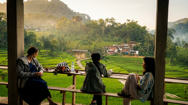 Three people relax on a wooden terrace overlooking lush green rice terraces, forested hills, and a village. The scene is tranquil, with soft morning light.