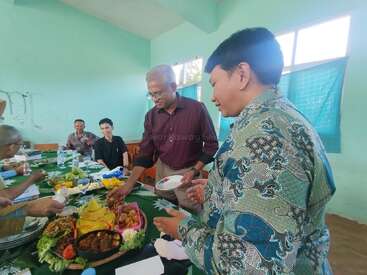 A group of people are gathered around a table sharing a meal in a brightly lit room, engaging in conversation and serving traditional Indonesian food together.