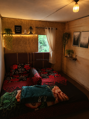 A cozy bedroom with a double bed, warm lighting, plants, woven walls, framed art, a curtain window, blanket, and minimalist decor, creating a peaceful atmosphere.