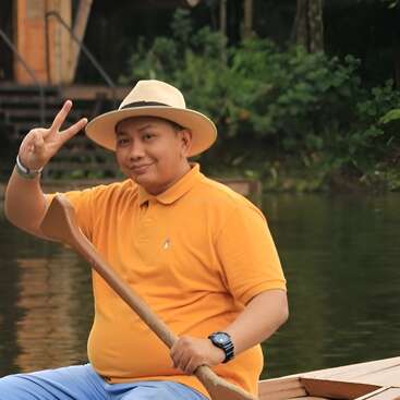 A smiling man wearing a yellow shirt and a hat sits in a boat, holding a paddle and flashing a peace sign by a calm river.
