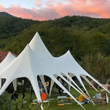 As pessoas relaxam sob uma tenda branca bem diferente, num espaço ao ar livre coberto de grama, cercado por árvores e montanhas ao pôr do sol, com tapetes de ioga e almofadas espalhados por todo o lado.