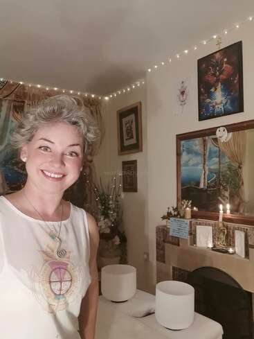 The image depicts a woman standing in a room with white walls, adorned with artwork and string lights, featuring a massage table with two white bowls in the foreground.