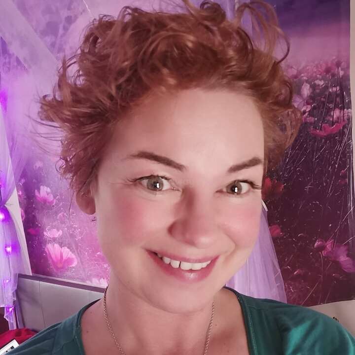 The image shows a woman with short red hair, wearing a green shirt and a necklace, in front of a floral-patterned wall with purple lights and curtains.