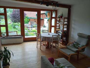Uma sala de estar aconchegante com piso de madeira, janelas grandes com vista para o jardim, mesa de jantar redonda com cadeiras, poltrona, sofá, estante de livros e plantas de interior criam um ambiente acolhedor.