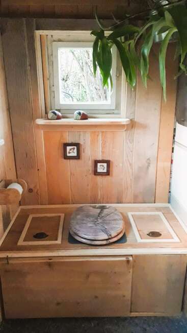 The image depicts a wooden toilet with a window above it, featuring a plant hanging from the ceiling and two small pictures on the wall.