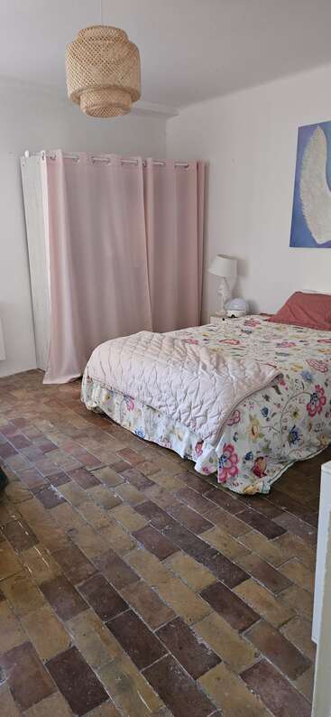 Une chambre à coucher confortable avec une literie florale, un édredon, des rideaux roses, une lampe de chevet, un plafonnier tressé, des œuvres d'art au mur et un sol en briques rustiques. Atmosphère paisible.