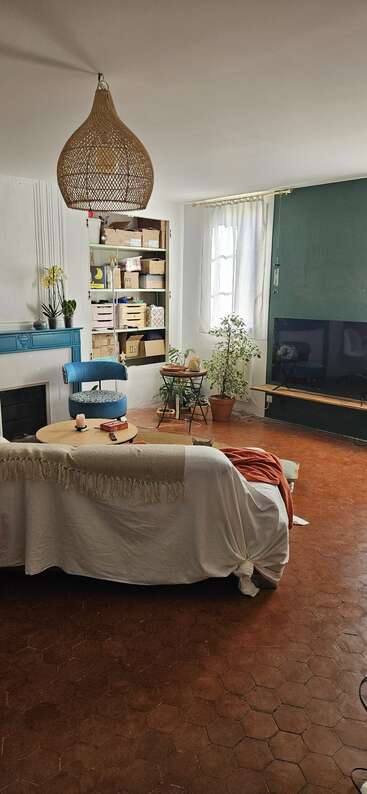 Salon confortable avec sol en terre cuite, canapé blanc, fauteuil bleu, plantes en pot, bibliothèque, mur d'accent vert, télévision, table basse, lampe en osier et lumière du soleil.